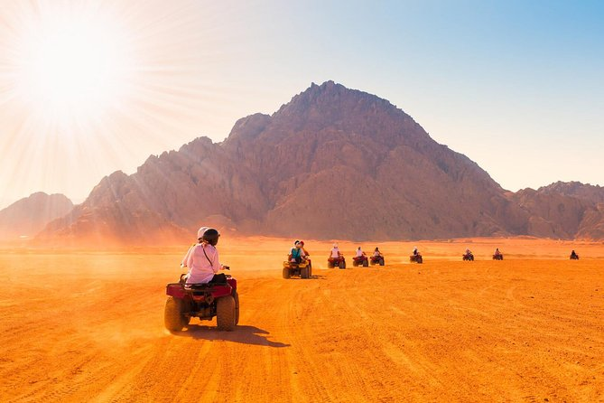 Travelers riding ATVs across golden desert sand toward rugged mountains