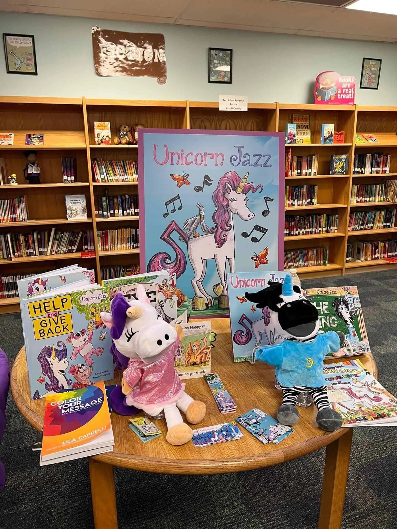 Unicorn Jazz books, a display sign and unicorn and zebra puppets arranged on a table in front of library shelves.