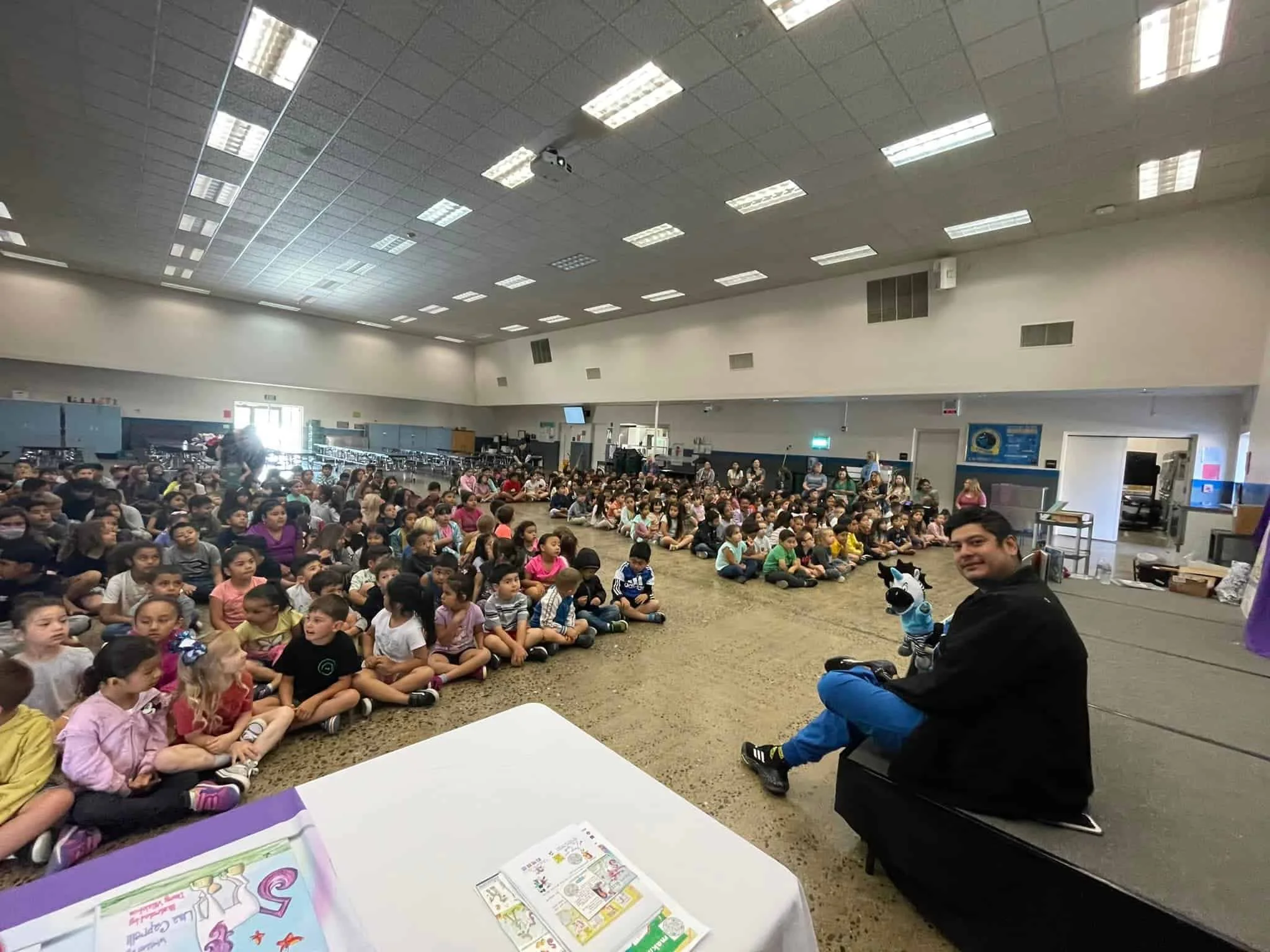 Children seated in a large room with a presenter holding a zebra puppet.
