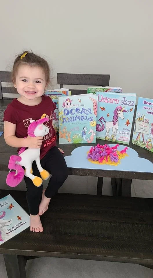 Child holding a unicorn toy beside Unicorn Jazz books and a colorful craft project.