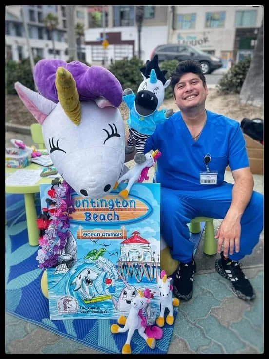 An adult in blue scrubs holding a zebra puppet beside a Unicorn Jazz book and plush display outdoors.