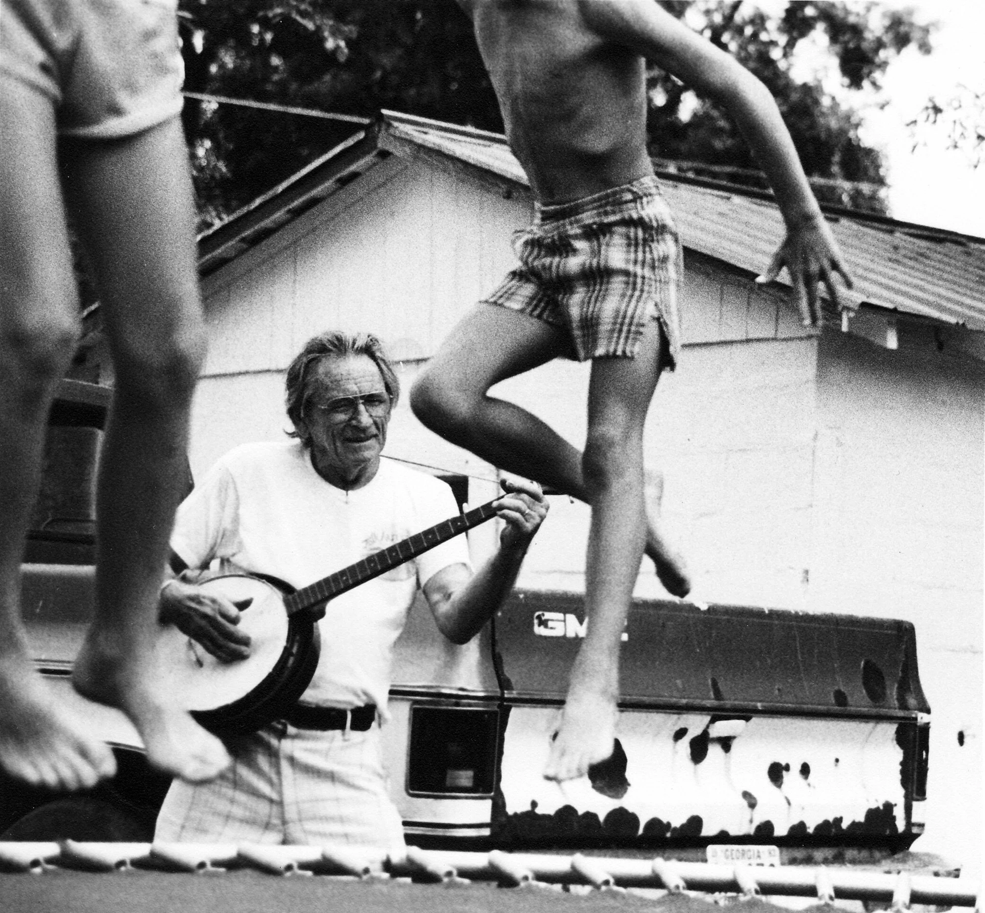 Howard Finster playing banjo while children jump on a trampoline at Paradise Garden in 1984