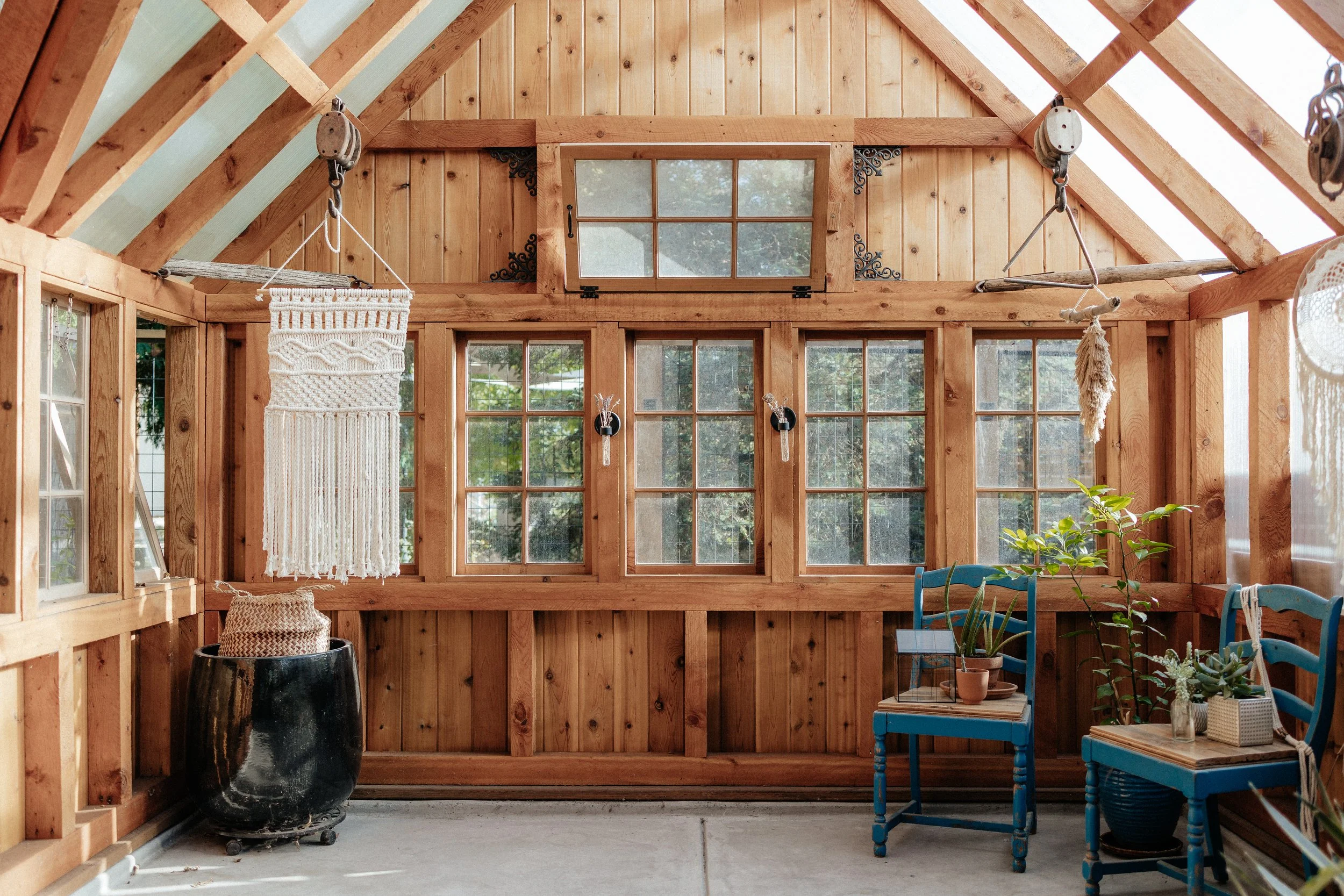 Sunlit cedar greenhouse with vintage windows, macrame, blue chairs and potted plants