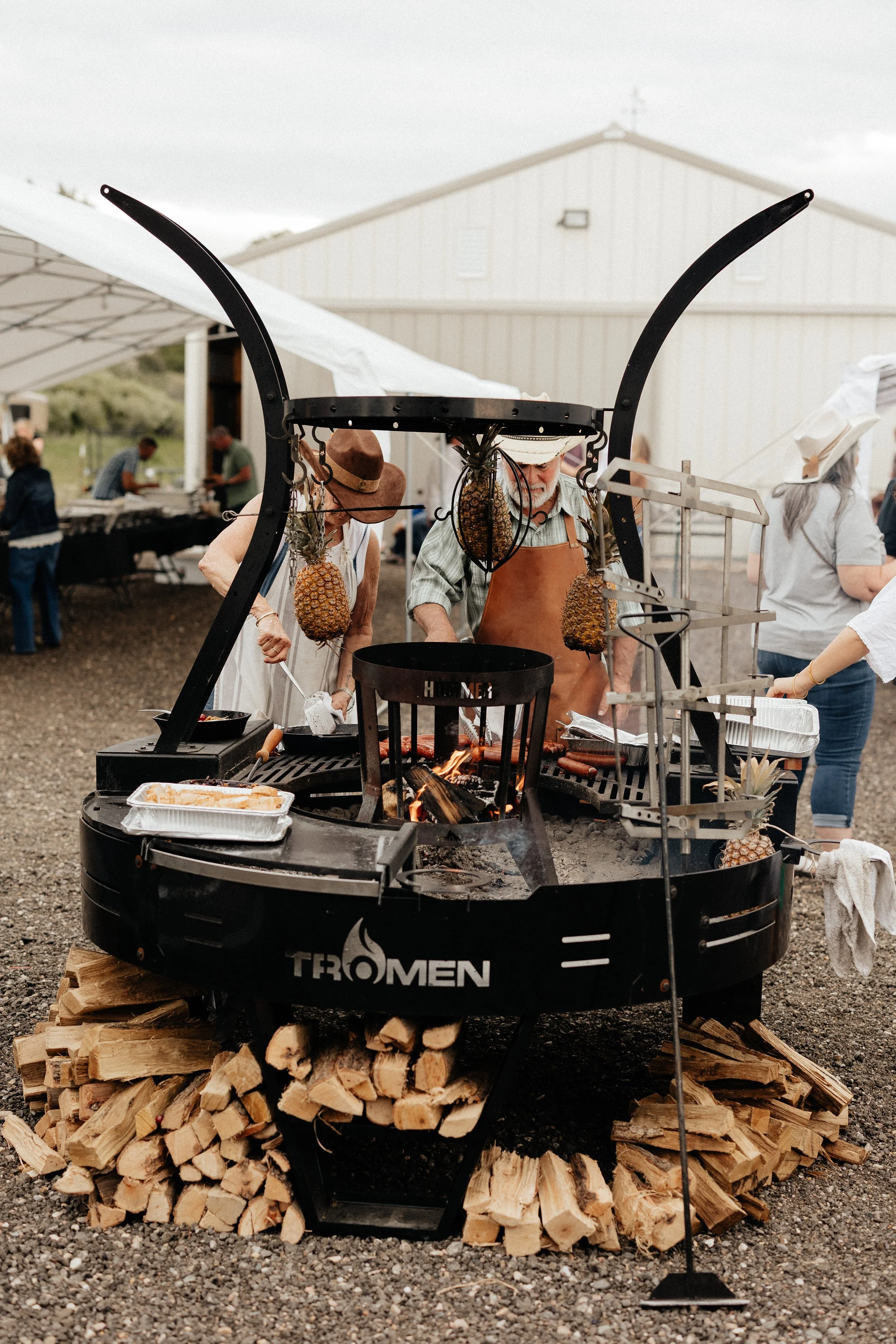 Cooks preparing dinner over an open wood-fired grill with hanging pineapples