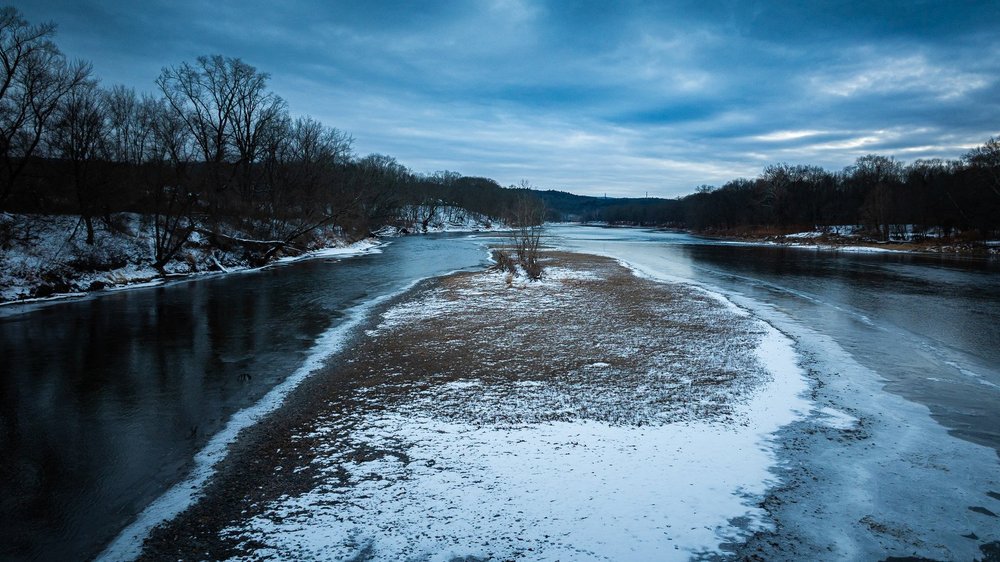 Snow edges a river sandbar beneath bare trees and a blue winter sky.