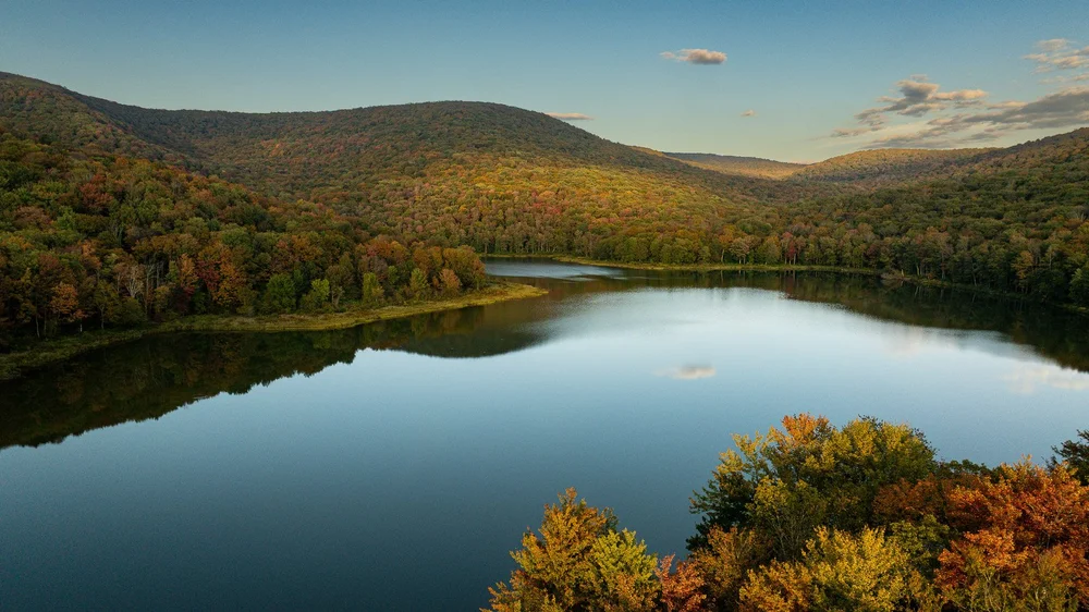 A still lake reflects wooded hills as the foliage begins to turn gold.