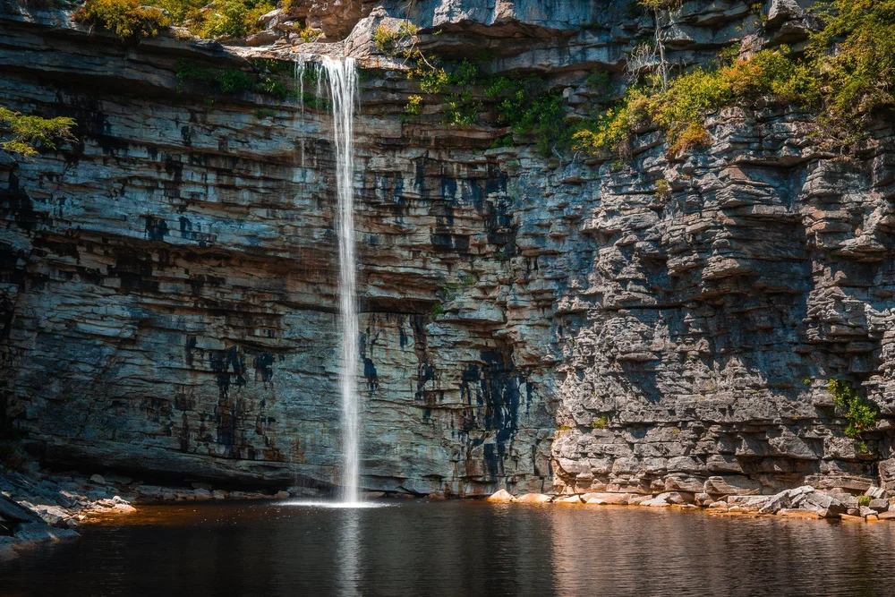 A slender waterfall drops down a tall, layered cliff into a still pool.