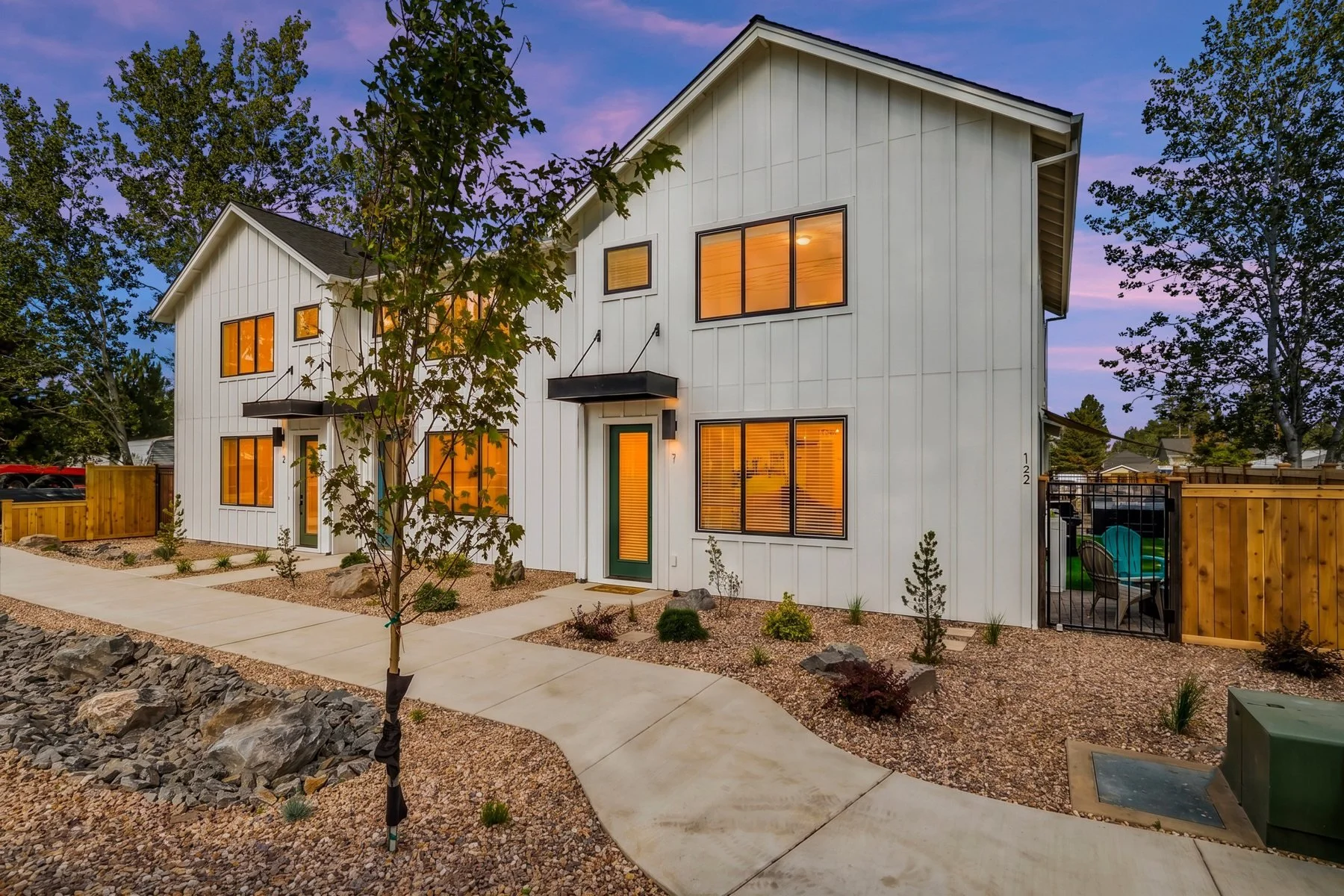 Locust, white board-and-batten multi-family townhomes in Sisters at dusk with lit windows and desert landscaping