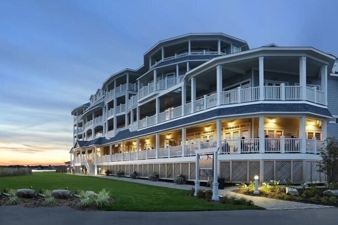 The Madison Beach Hotel at dusk, a large shingled building with wraparound porches and lit windows, under a warm sunset sky.