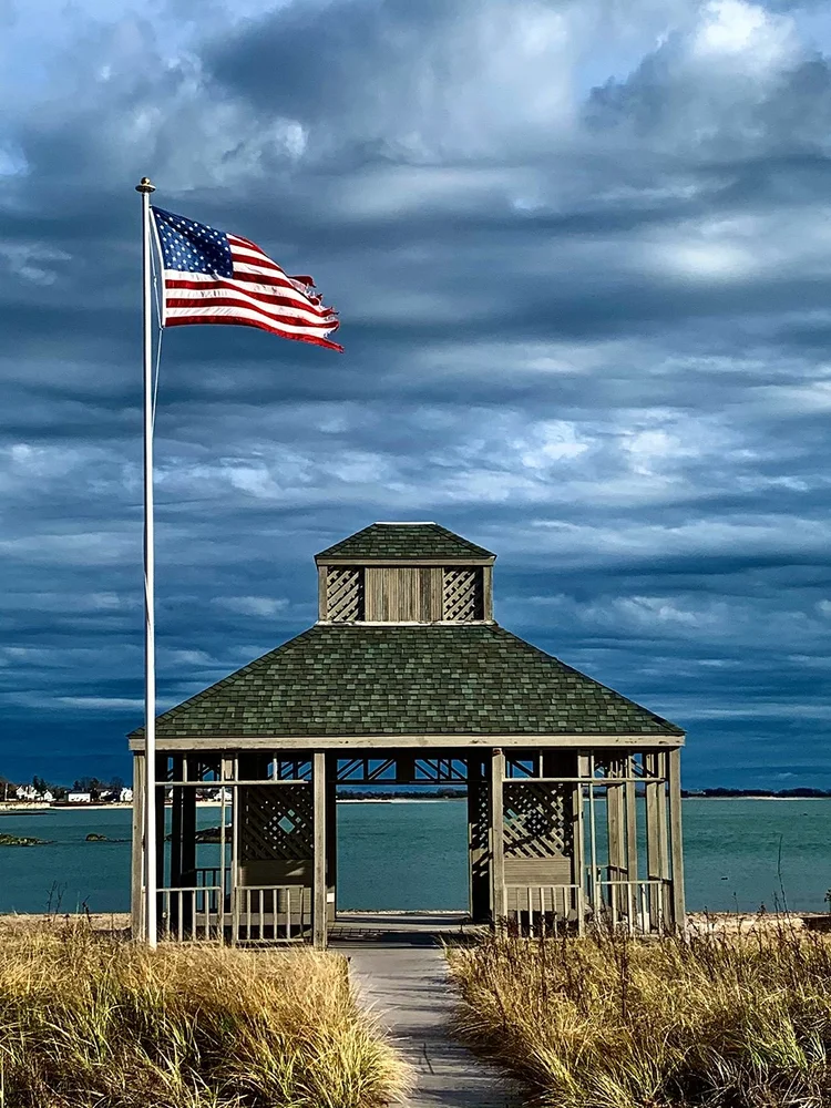 A shingled beach pavilion flying an American flag on the Madison shoreline, under a big cloudy sky.
