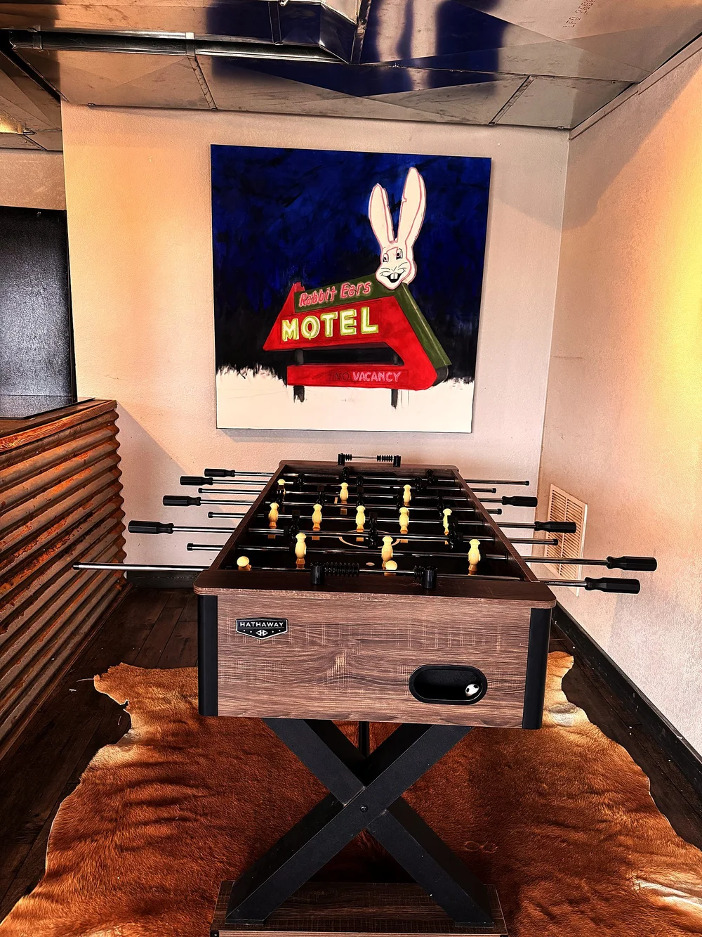 A foosball table on a cowhide rug beneath a painting of a rabbit motel sign