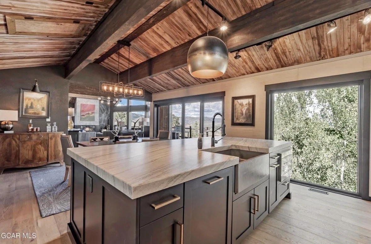 A stone kitchen island with a farmhouse sink beside floor-to-ceiling windows into the aspens