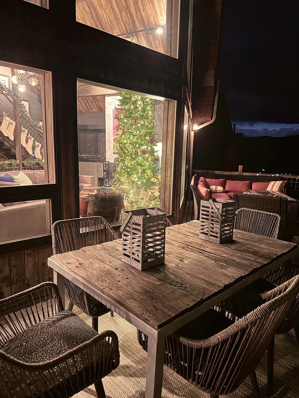 The deck at night with string lights, lanterns on the table and the tree glowing through the windows