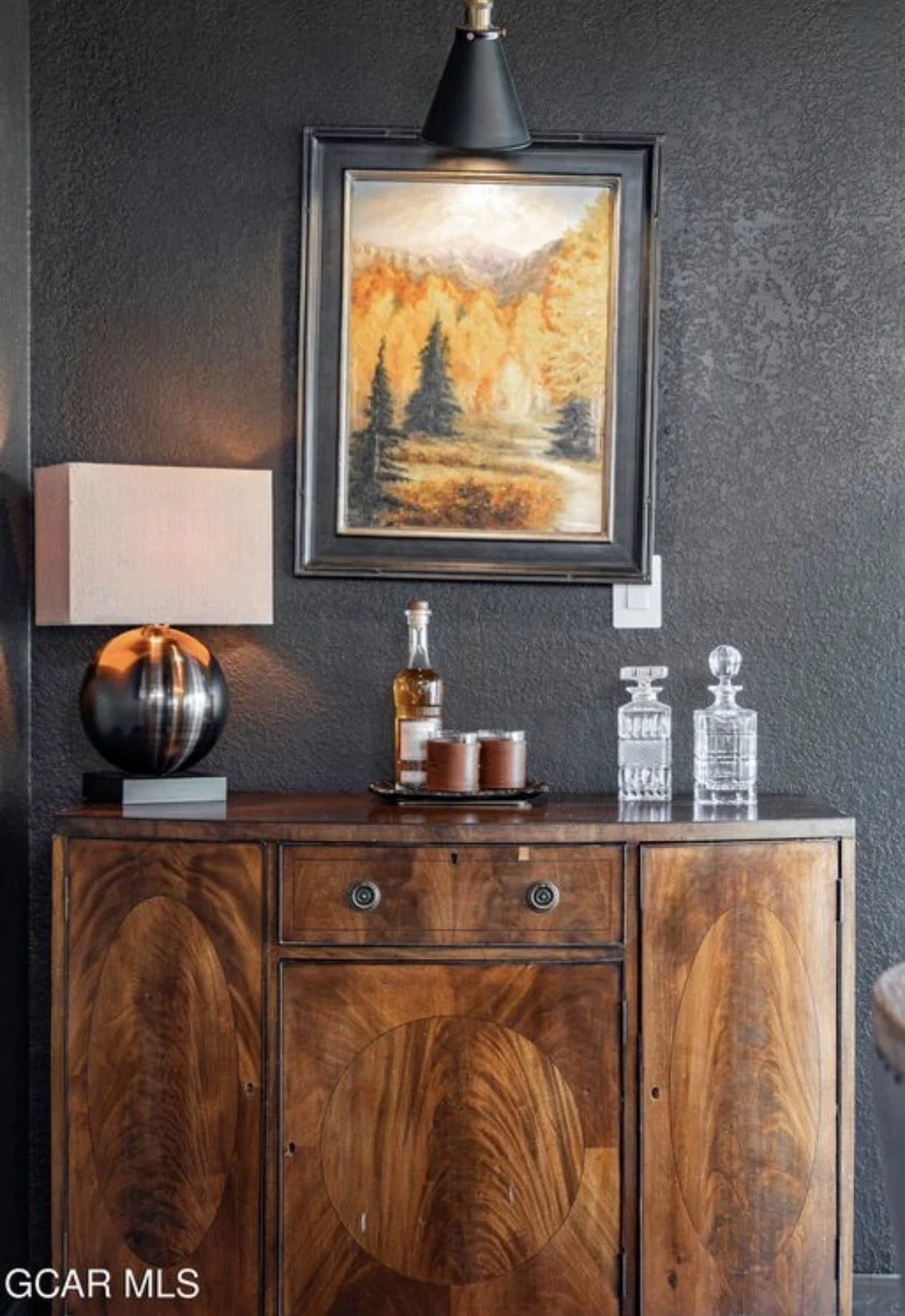 An oil painting of autumn aspens above a walnut sideboard with crystal decanters