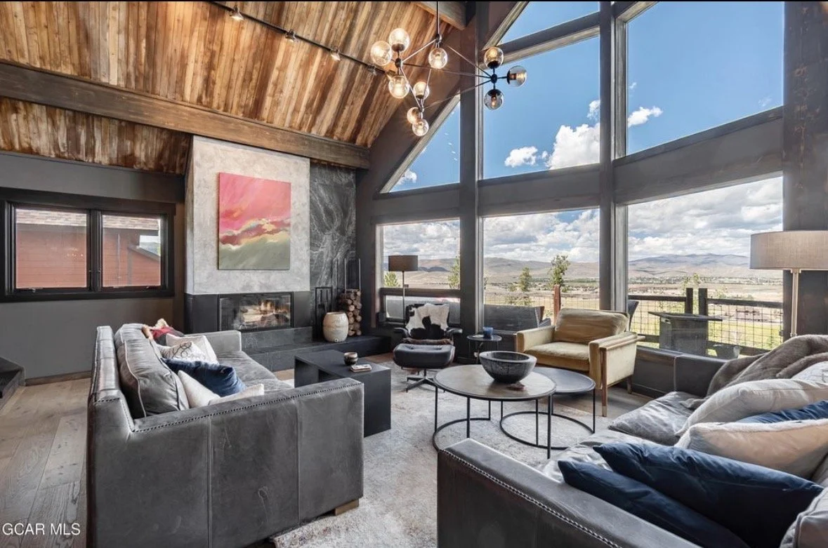 The great room with leather sofas, a marble-clad fireplace, a pink abstract painting and two-story windows onto the valley