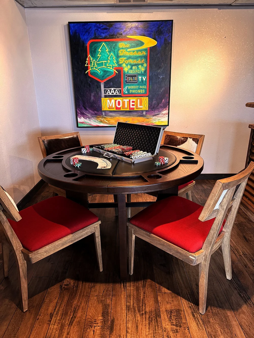 A round poker table with red chairs beneath a painting of a vintage motel sign