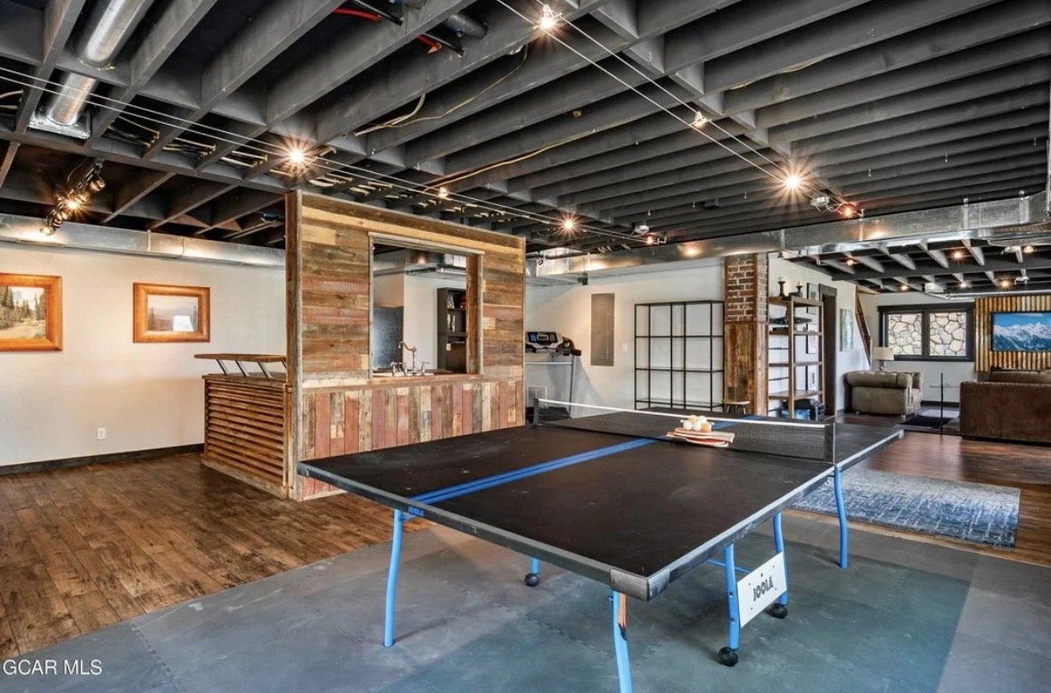 The lower-level game room with a ping pong table, a reclaimed-wood wet bar and glass doors to the aspens