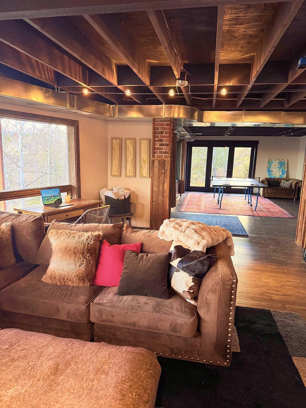 The lower-level lounge with a deep sectional, a desk by the windows, and the game area beyond