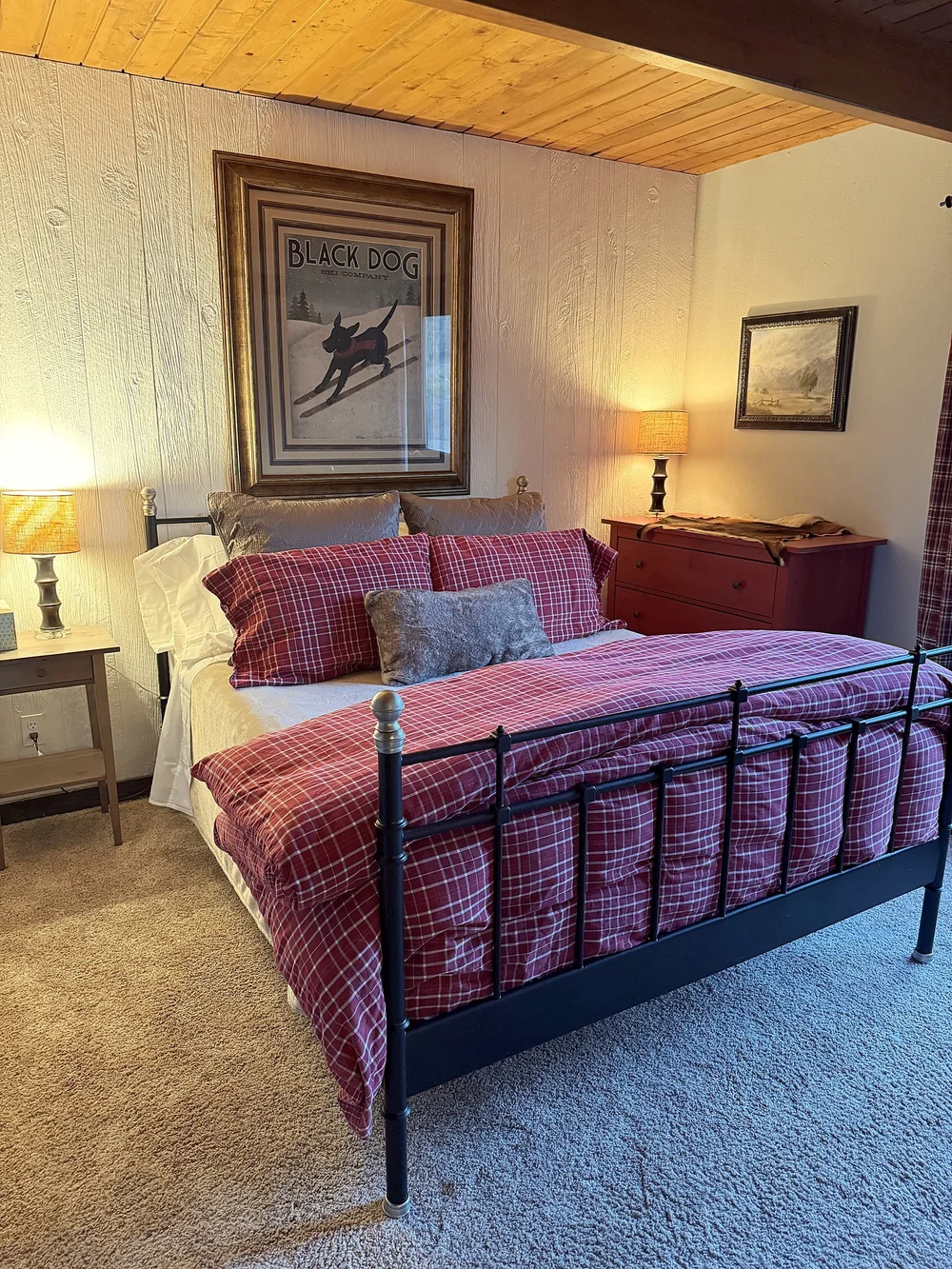A queen bedroom with an iron bed, red plaid bedding and a vintage Black Dog ski poster