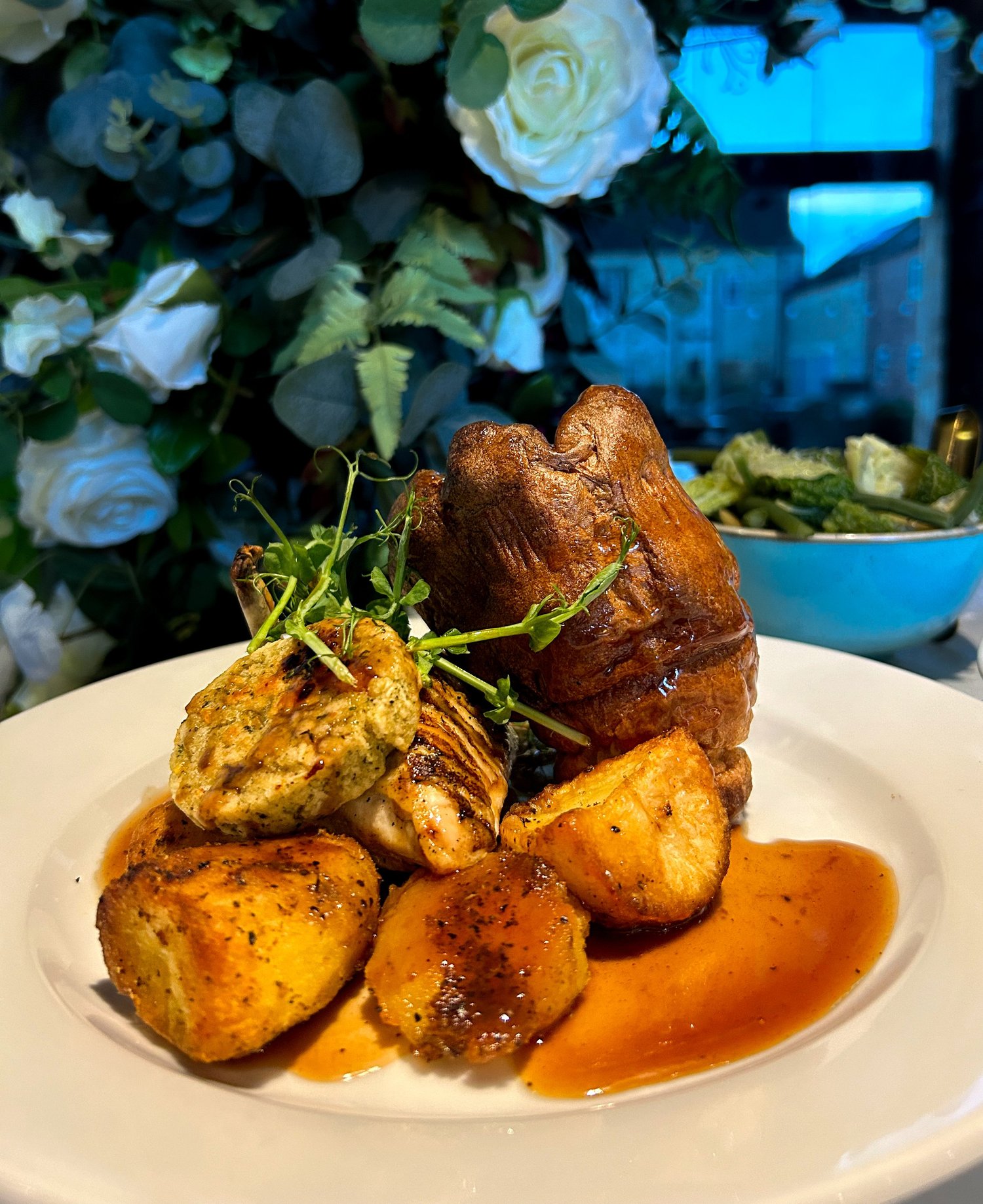 A roast dinner of Yorkshire pudding, golden roast potatoes and gravy on a white plate, with flowers behind