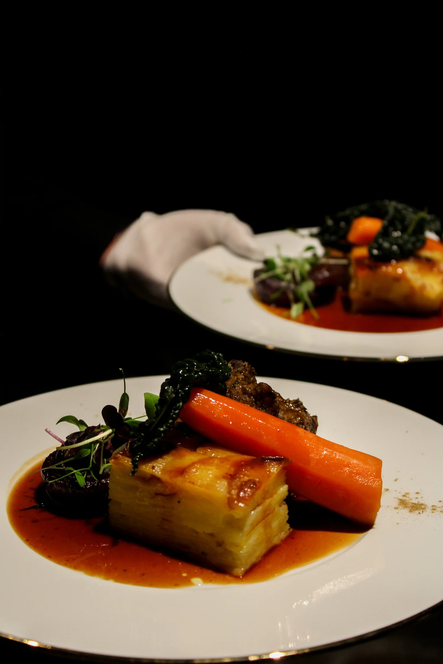 A plated beef main with carrot, kale and layered potato being served by a white-gloved waiter at an evening reception