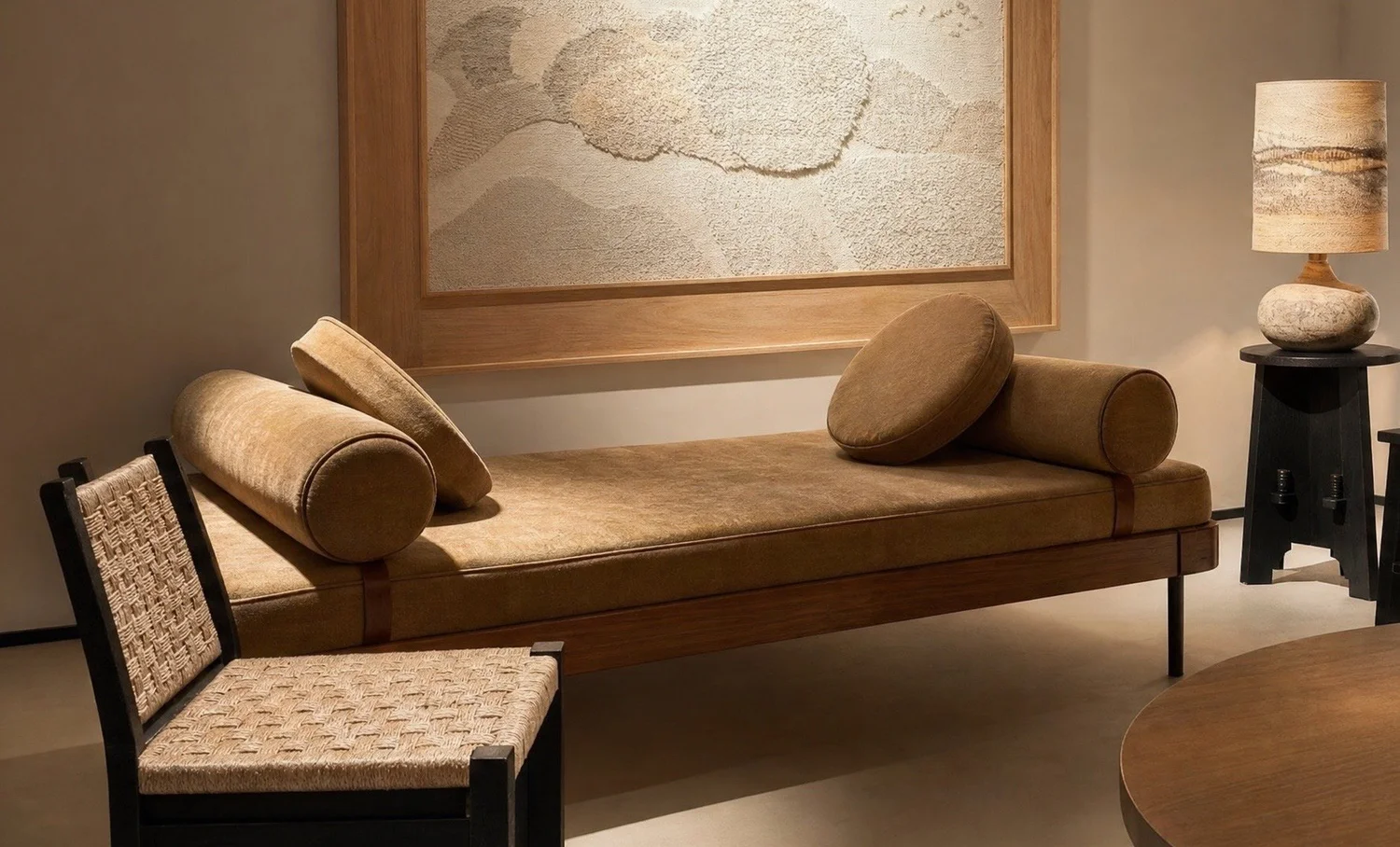 A daybed and woven chair beneath a textured artwork, lit by a stone lamp