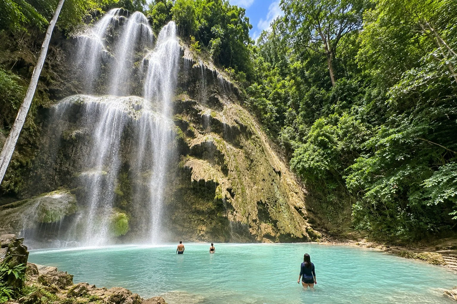 Visitors in a shallow turquoise pool beneath the broad cascade of Tumalog Falls