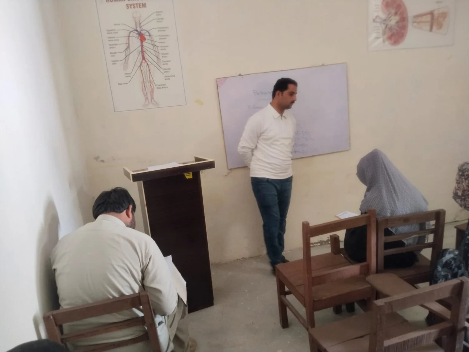 An instructor teaches human anatomy to paramedical diploma students in front of circulatory and renal system posters