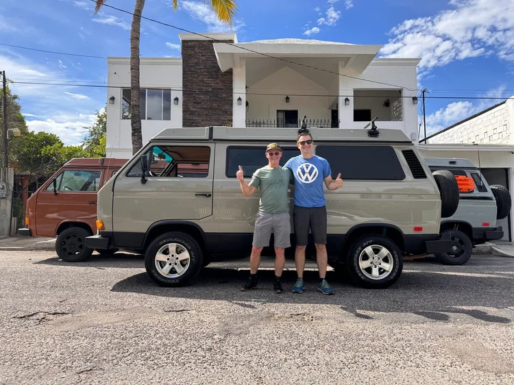 Two people beside a silver Volkswagen Vanagon outside a house