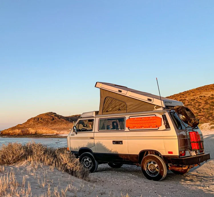 Beige Volkswagen Westfalia with its pop-top raised beside the coast