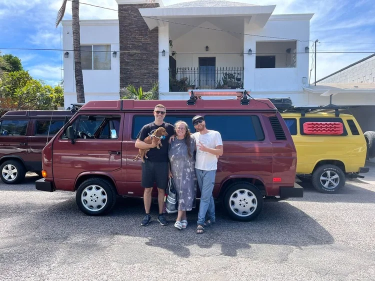 Three people beside a burgundy Volkswagen camper outside the workshop