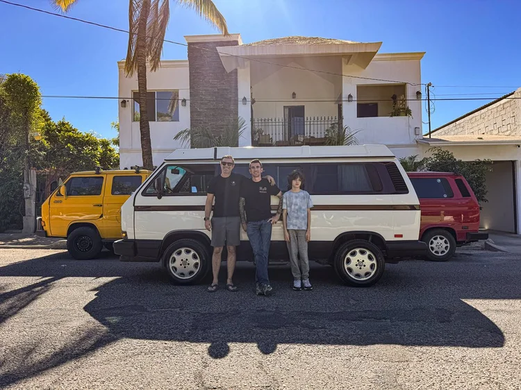 Three people beside a white Volkswagen camper outside the workshop