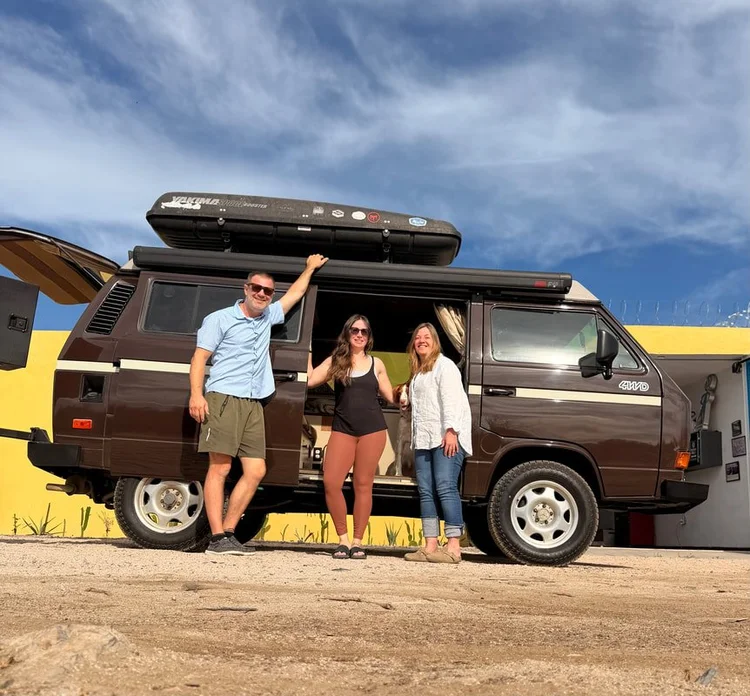 Three people beside a brown Volkswagen camper with roof cargo