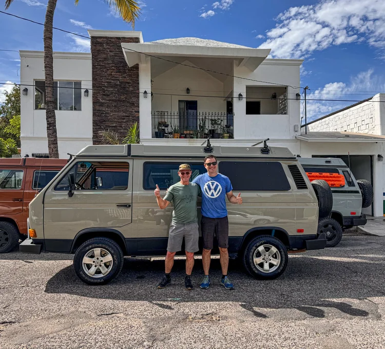 Two people beside a gray Volkswagen camper outside the workshop