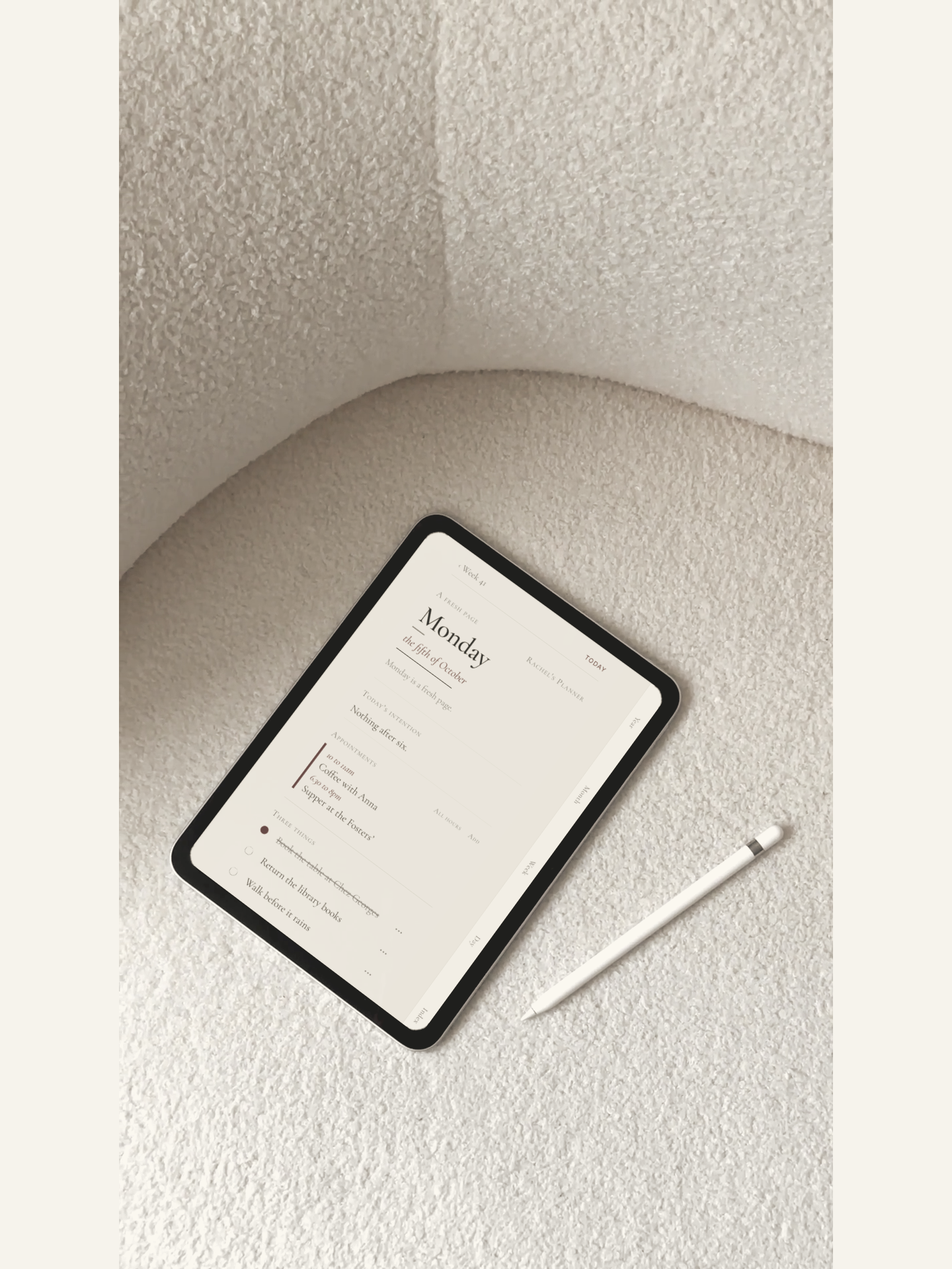 An iPad on a bouclé chair showing a Monday page in Lundi, the Pencil beside it