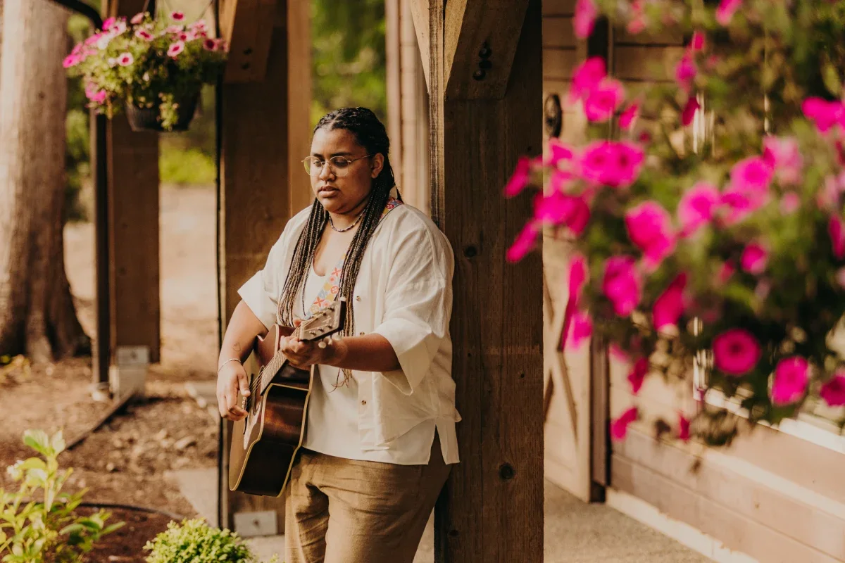 Mye playing acoustic guitar on a porch beside pink flowers