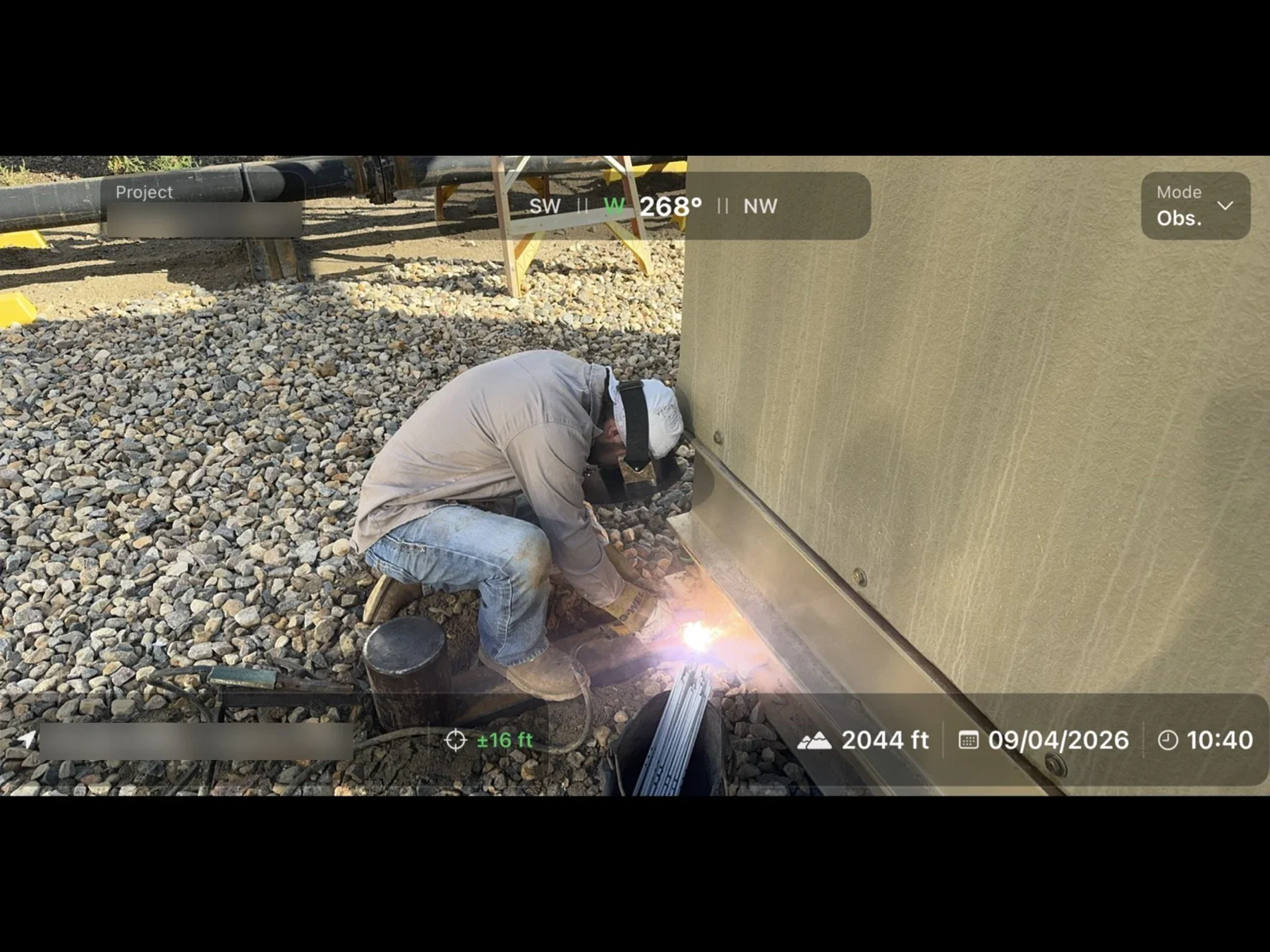 Welder working at the base of an equipment shelter, with location, heading, date and time stamped on the photo