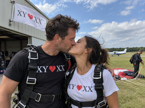 Couple in skydiving harnesses kissing after landing beside an airfield hangar.
