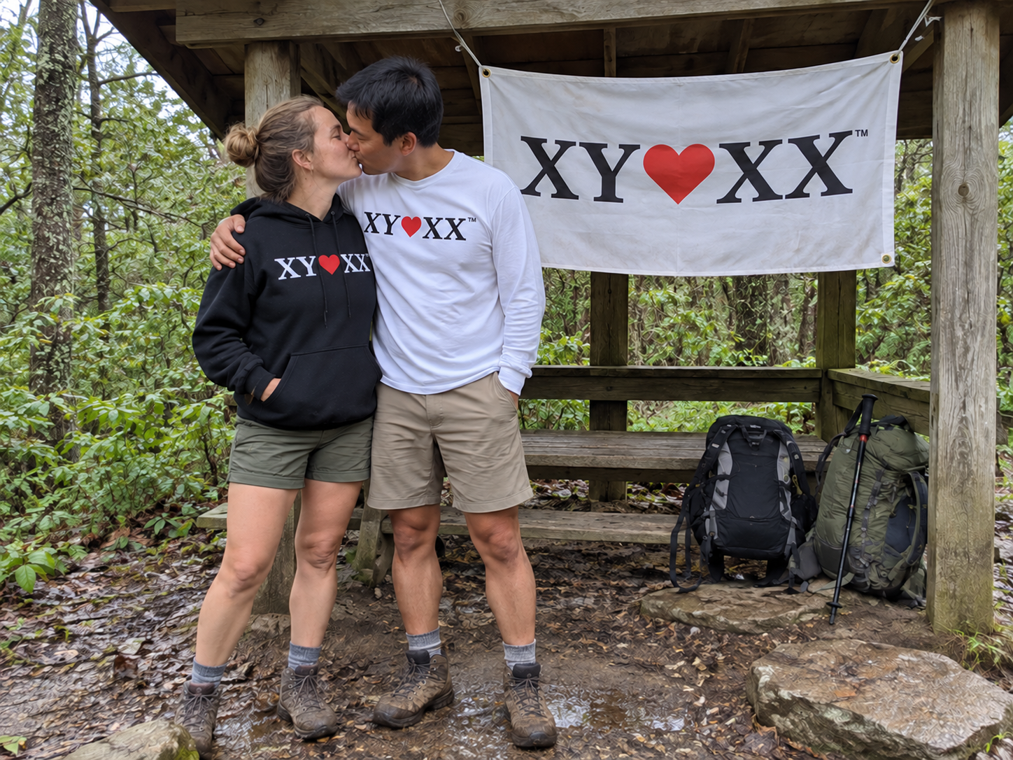 Couple in a hoodie and long-sleeve shirt kissing beneath a flag tied to a woodland shelter.