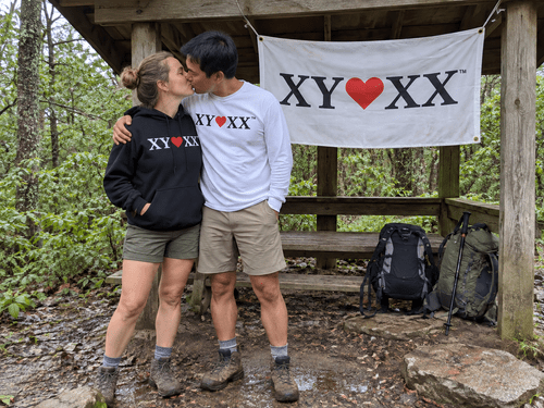 Couple in a hoodie and long-sleeve shirt kissing beneath a flag tied to a woodland shelter.