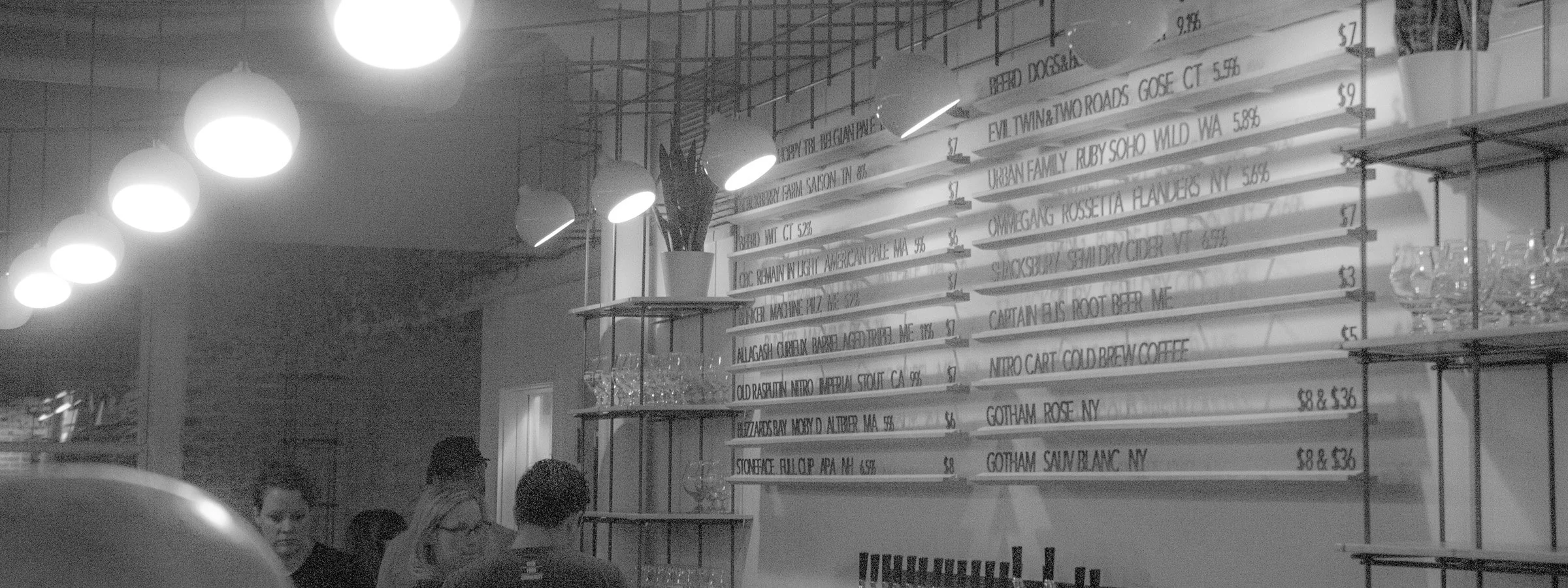 A row of globe pendants lit above a bar, beside a wall of hand-lettered boards listing what is on tap.