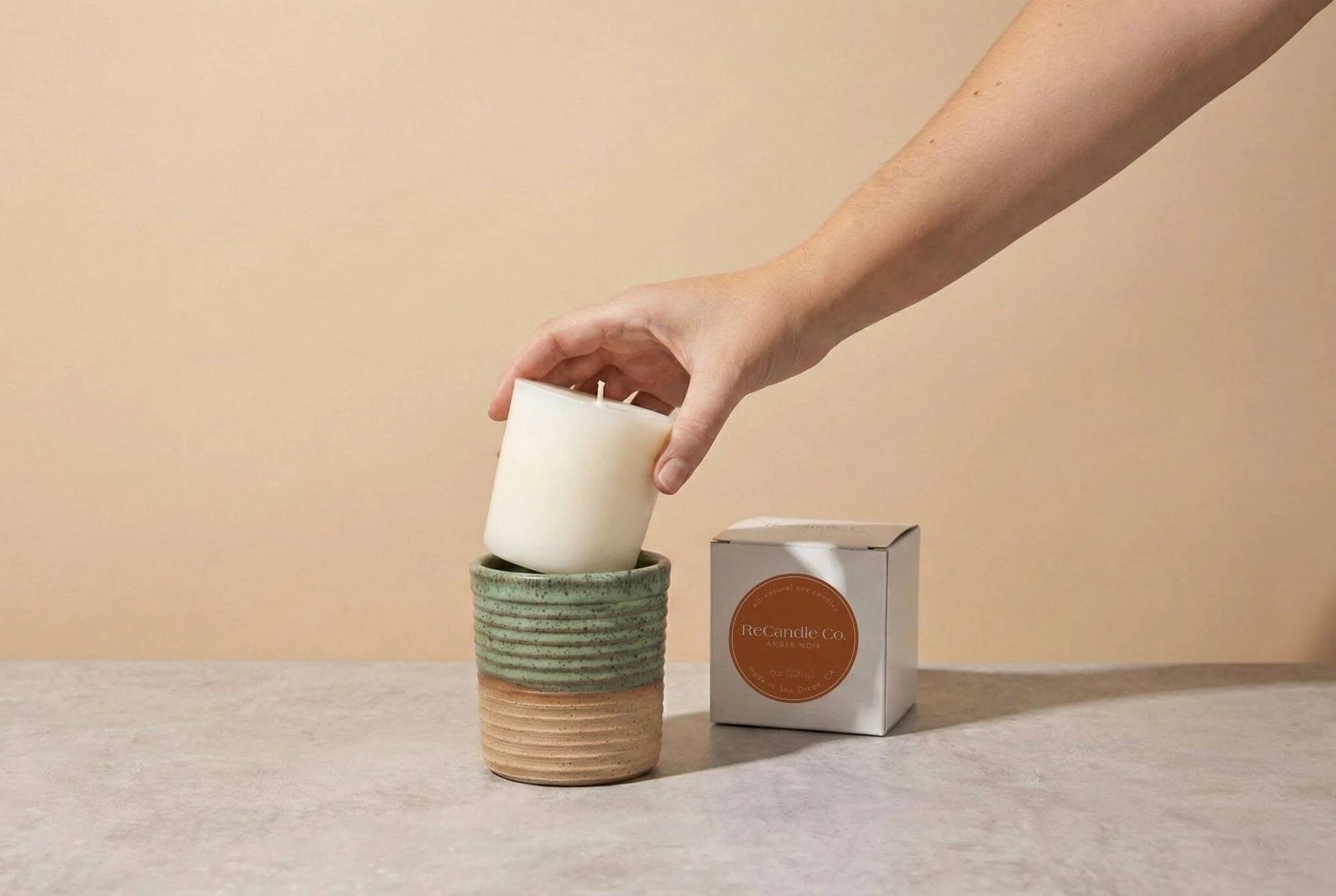 A candle refill being placed into a handmade green ceramic vessel beside ReCandle packaging