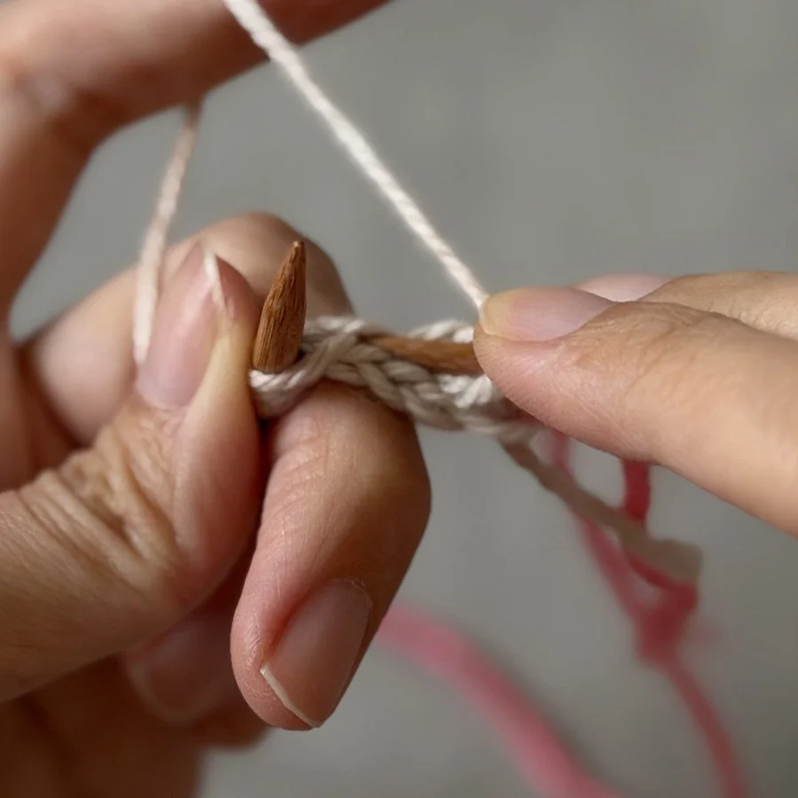 Close-up of the reverse-side strand being lifted onto the left needle beside the white centre stitch.