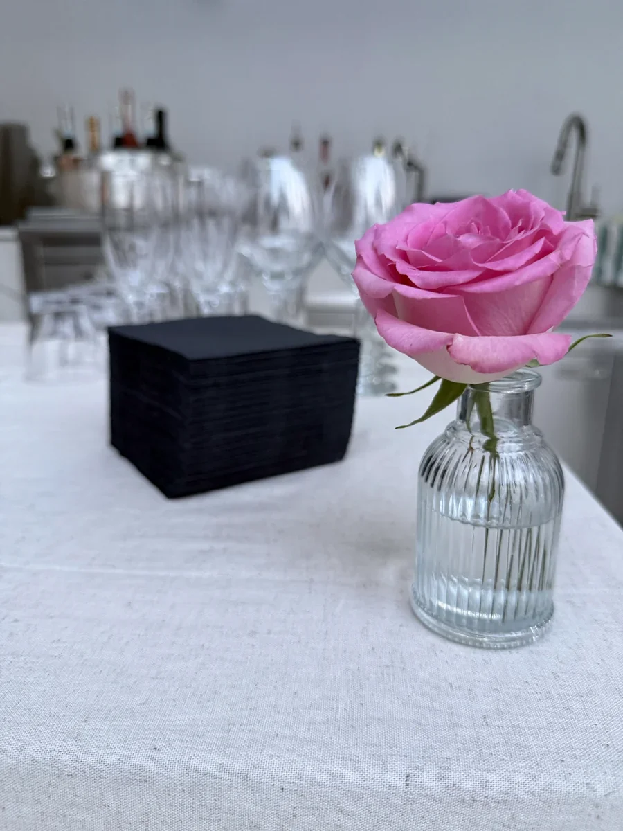 A pink rose beside arranged glassware and black cocktail napkins.