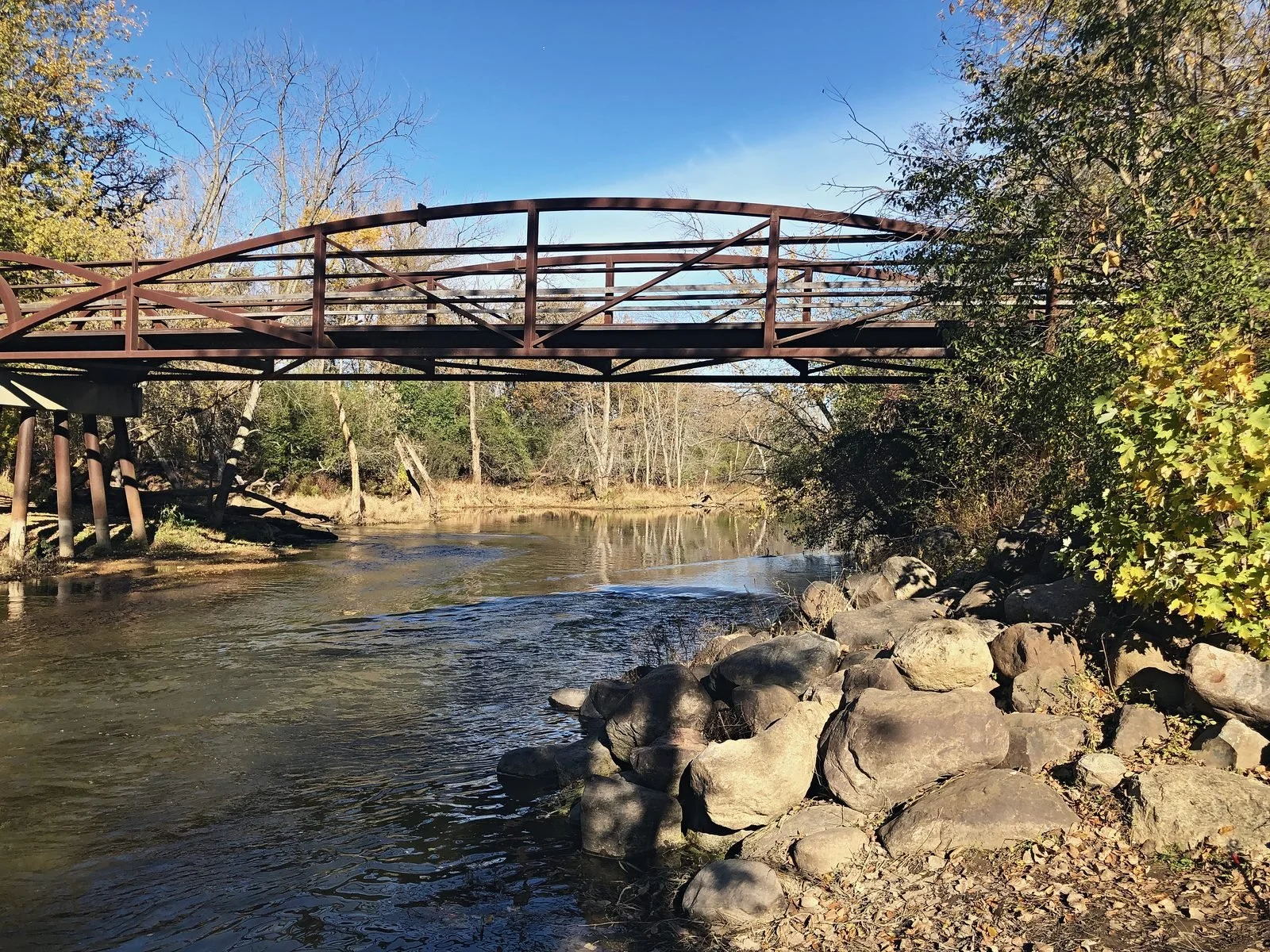 Rusted steel truss footbridge over a shallow river lined with boulders in autumn