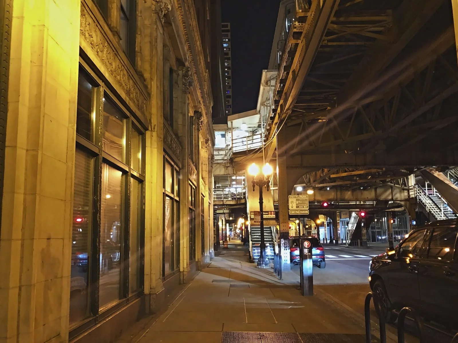 Sidewalk under the elevated train tracks at night, lit by a vintage street lamp