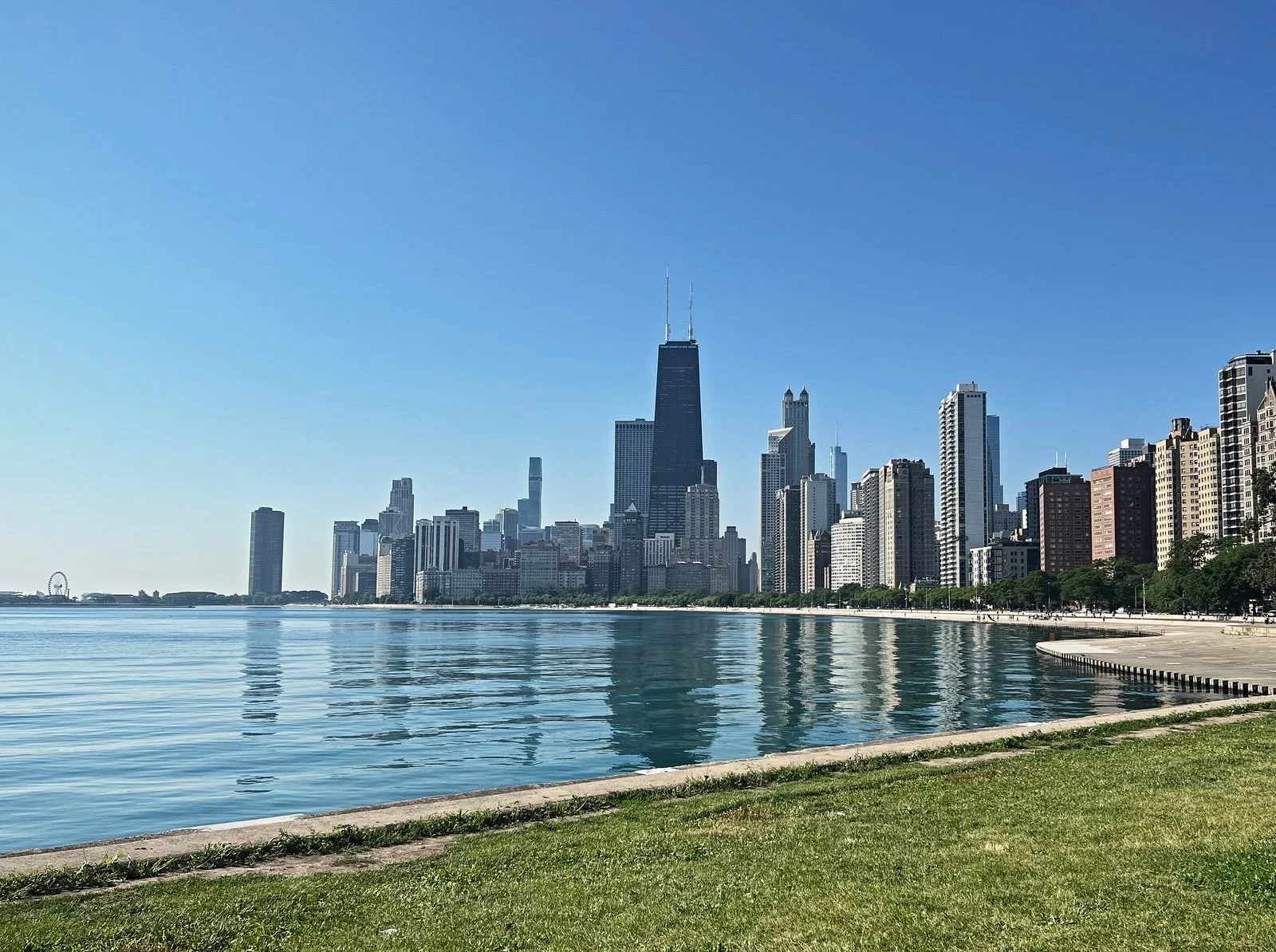 The Chicago skyline across the calm water of Lake Michigan, seen from a grassy lakefront park on a clear summer morning