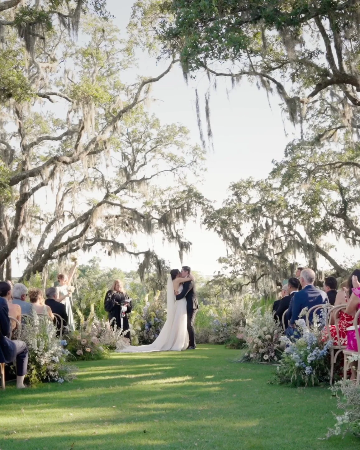 Jenny and Garrett kissing beneath live oaks and Spanish moss at The Dunlin, framed by wedding flowers and guests