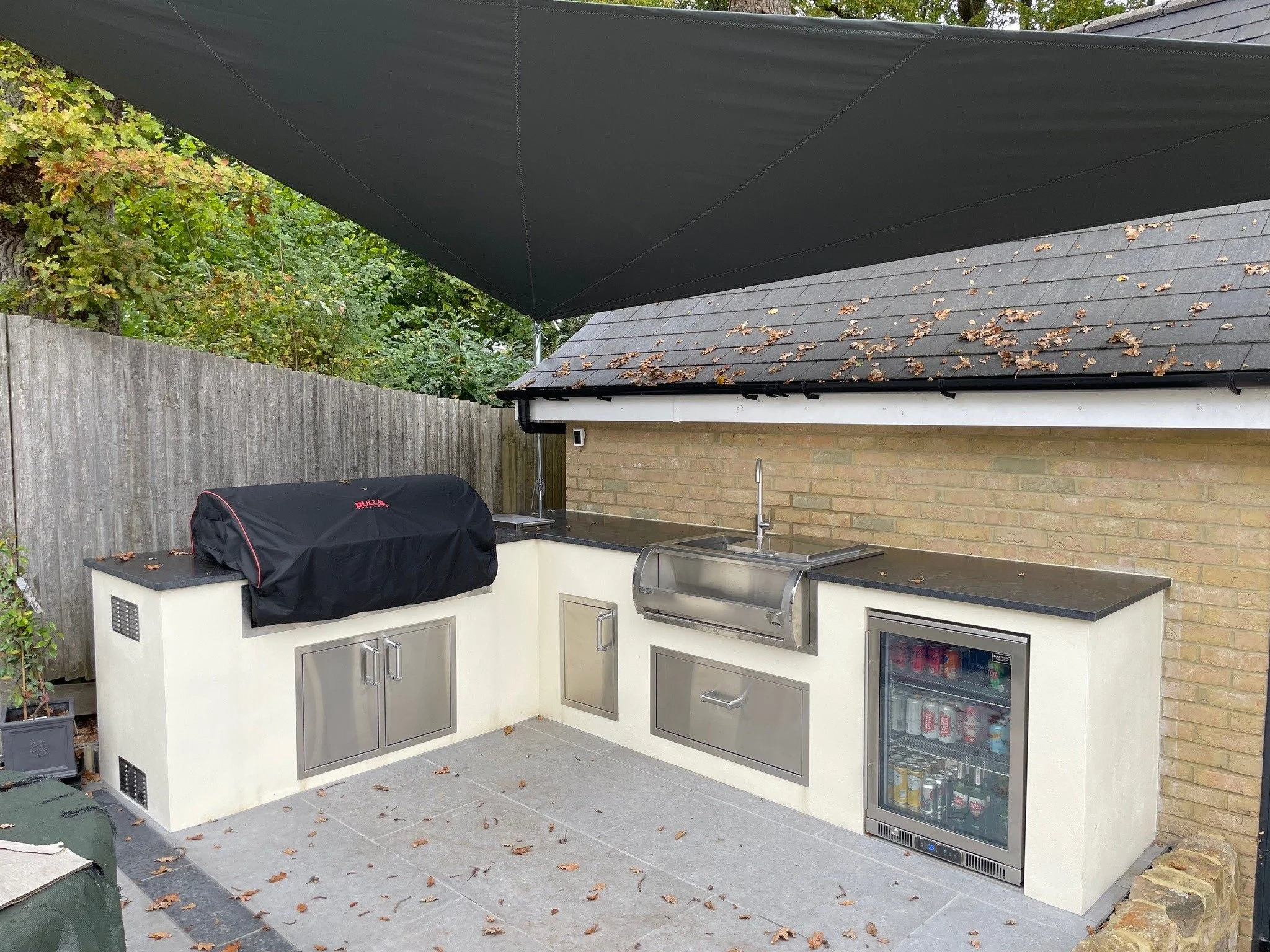 Installed outdoor kitchen beneath a sail shade, with a covered barbecue, sink, storage, worktops and drinks fridge.