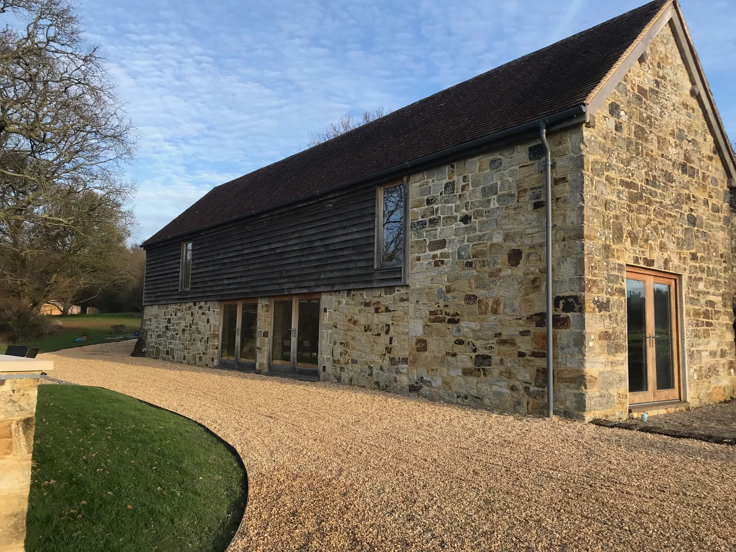 Stonework and timber cladding at Dewlands Barn, framed by lawn and a curved gravel approach.
